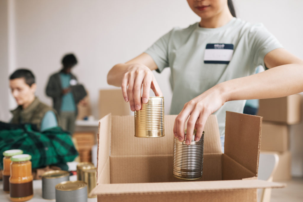 Close up of young woman packing canned food to boxes at charity and donations event, copy space